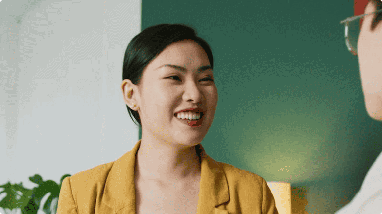 Seorang wanita dengan jaket kuning berdiri, sedang berbicara dengan seseorang di lingkungan yang nyaman, dikelilingi dinding hijau dan tanaman hias.