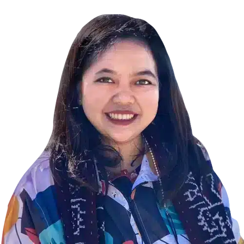 Seorang wanita mengenakan pakaian berwarna-warni sambil berdiri dengan latar belakang hitam. Dia tampak tersenyum, menciptakan suasana yang ramah.