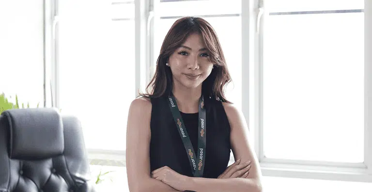 Seorang wanita berdiri dengan percaya diri, mengenakan gaun hitam dan lanyard, di dalam ruangan terang dengan jendela besar di latar belakang.