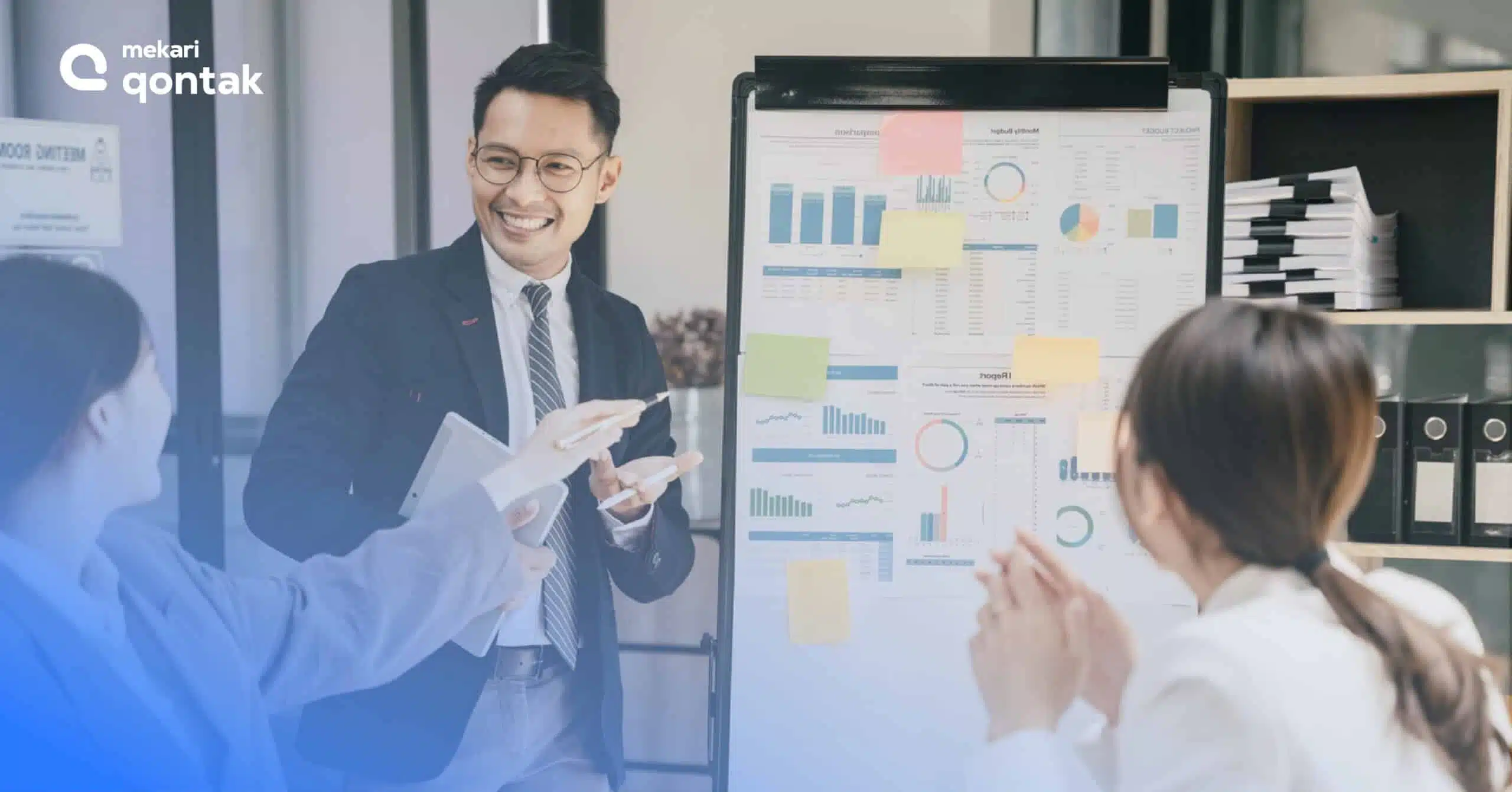 Seorang presenter menjelaskan data dan grafik pada papan tulis sementara rekan-rekannya memperhatikan dengan antusias di ruangan rapat modern.