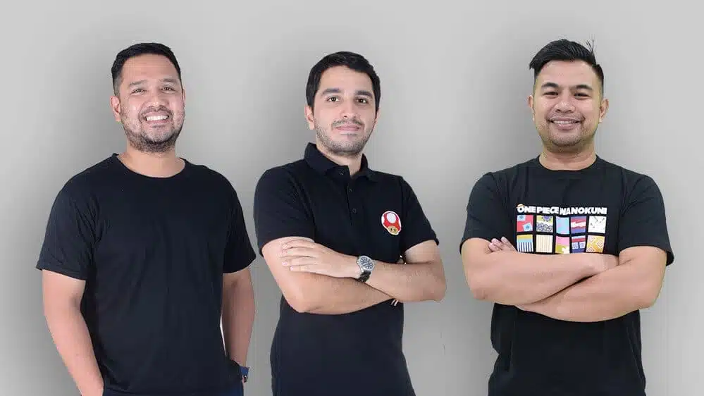 Tiga pria berdiri berjejer, mengenakan kaos hitam yang berbeda, dengan latar belakang abu-abu polos. Mereka menunjukkan sikap percaya diri.