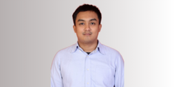 Pria mengenakan kemeja biru berdiri tegak di depan latar belakang abu-abu yang halus, memberikan kesan formal dan profesional.