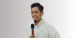 Seseorang berbaju putih memegang mikrofon berwarna merah dan biru, berdiri di depan latar belakang yang lembut. Ia siap berbicara di depan audiens.
