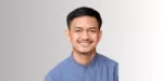 Seorang pria mengenakan kemeja biru dengan kerah, berdiri di depan latar belakang gradasi warna netral. Dia terlihat tenang dan fokus.