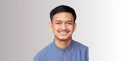 Seorang pria mengenakan kemeja biru dengan kerah, berdiri di depan latar belakang gradasi warna netral. Dia terlihat tenang dan fokus.