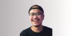 Seorang pria mengenakan kaos hitam dan topi, berdiri dengan latar belakang yang berwarna gradasi lembut, terlihat percaya diri dan santai.