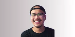 Seorang pria mengenakan kaos hitam dan topi, berdiri dengan latar belakang yang berwarna gradasi lembut, terlihat percaya diri dan santai.