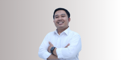 Seseorang mengenakan kemeja putih dengan sikap percaya diri, berdiri dengan tangan disilangkan di depan tubuhnya, di latar belakang yang bergradasi halus.