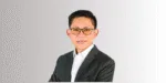 Seseorang mengenakan jas gelap dan kemeja putih, berdiri dengan pose percaya diri di latar belakang berwarna netral, menciptakan kesan profesional.