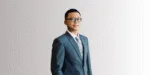 Pria mengenakan jas formal berwarna biru, berdiri tegak di depan latar belakang gradasi abu-abu, menciptakan kesan profesional dan elegan.