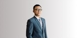 Pria mengenakan jas formal berwarna biru, berdiri tegak di depan latar belakang gradasi abu-abu, menciptakan kesan profesional dan elegan.