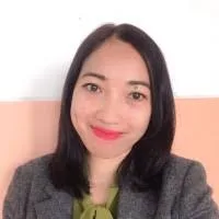 Seorang wanita dengan rambut panjang dan hitam mengenakan jas abu-abu, berdiri di depan dinding berwarna pastel. Dia tampak percaya diri dan profesional.