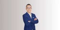 Seorang pria berpakaian jas biru, dengan tangan disilangkan di dada, berdiri dengan latar belakang sederhana berwarna putih. Dia menunjukkan sikap percaya diri.