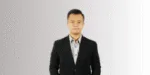 Pria berbalut jas formal berdiri tegak di latar belakang berwarna netral, memberikan kesan profesional dan siap untuk berinteraksi.