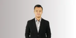 Pria berbalut jas formal berdiri tegak di latar belakang berwarna netral, memberikan kesan profesional dan siap untuk berinteraksi.