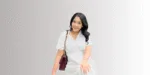 Seorang wanita berpakaian putih melangkah dengan percaya diri, mengulurkan tangan seolah menyapa atau mengajak interaksi, di latar belakang yang sederhana.
