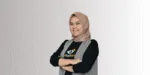 Seorang individu mengenakan jilbab beige dan vest dengan lengan dilipat, berdiri dengan sikap percaya diri di latar belakang berwarna gradasi.