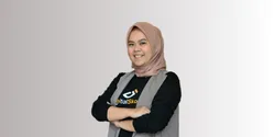 Seorang individu mengenakan jilbab beige dan vest dengan lengan dilipat, berdiri dengan sikap percaya diri di latar belakang berwarna gradasi.