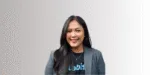 Seorang individu mengenakan jas formal dan kaos bertuliskan "Labis" sedang berdiri di depan latar belakang bergradasi yang lembut, menampilkan profesionalisme.