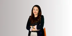 Seorang wanita berdiri dengan tangan disilangkan, mengenakan blazer hitam dan celana putih, tampak percaya diri di latar belakang yang terang dan minimalis.