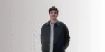 Seorang pria mengenakan jaket gelap dan kemeja yang menyenangkan berdiri dengan posisi santai, latar belakang halus yang cerah menambah kesan modern.