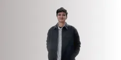 Seorang pria mengenakan jaket gelap dan kemeja yang menyenangkan berdiri dengan posisi santai, latar belakang halus yang cerah menambah kesan modern.