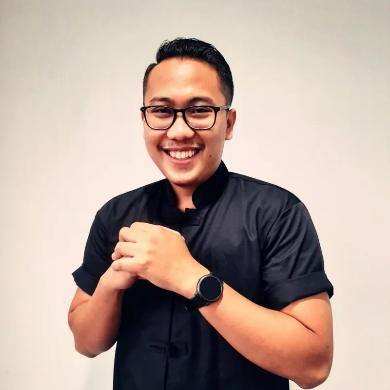 Seorang pria mengenakan kemeja hitam berdiri dengan tangan disilangkan di depan tubuhnya, berlatarkan dinding polos yang cerah.