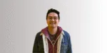 Seorang pria berdiri tegak mengenakan jaket denim dan sweater, dengan latar belakang sederhana dan terang, menciptakan suasana yang santai.
