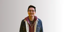 Seorang pria berdiri tegak mengenakan jaket denim dan sweater, dengan latar belakang sederhana dan terang, menciptakan suasana yang santai.