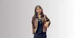 Seorang wanita mengenakan jaket cokelat dan rok hitam berdiri dengan percaya diri, dikelilingi oleh latar belakang yang netral dan sederhana.
