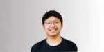 Seorang pria mengenakan kaos hitam berdiri dengan latar belakang gradasi yang cerah, memberikan kesan profesional dan fokus pada kehadirannya.