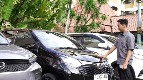 Sebuah mobil hitam terparkir di antara dua mobil lain, sementara seorang pria mengenakan seragam menghadap dan menyentuh kap mobil, siap membantu.